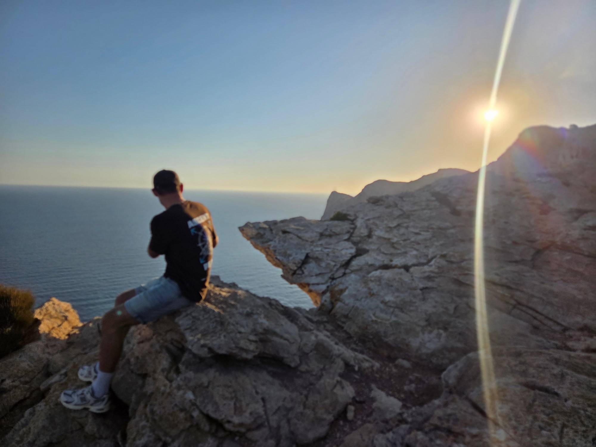 Someone sitting on a rocky cliff edge looking out over a calm sea, with the sun low over the ridge.