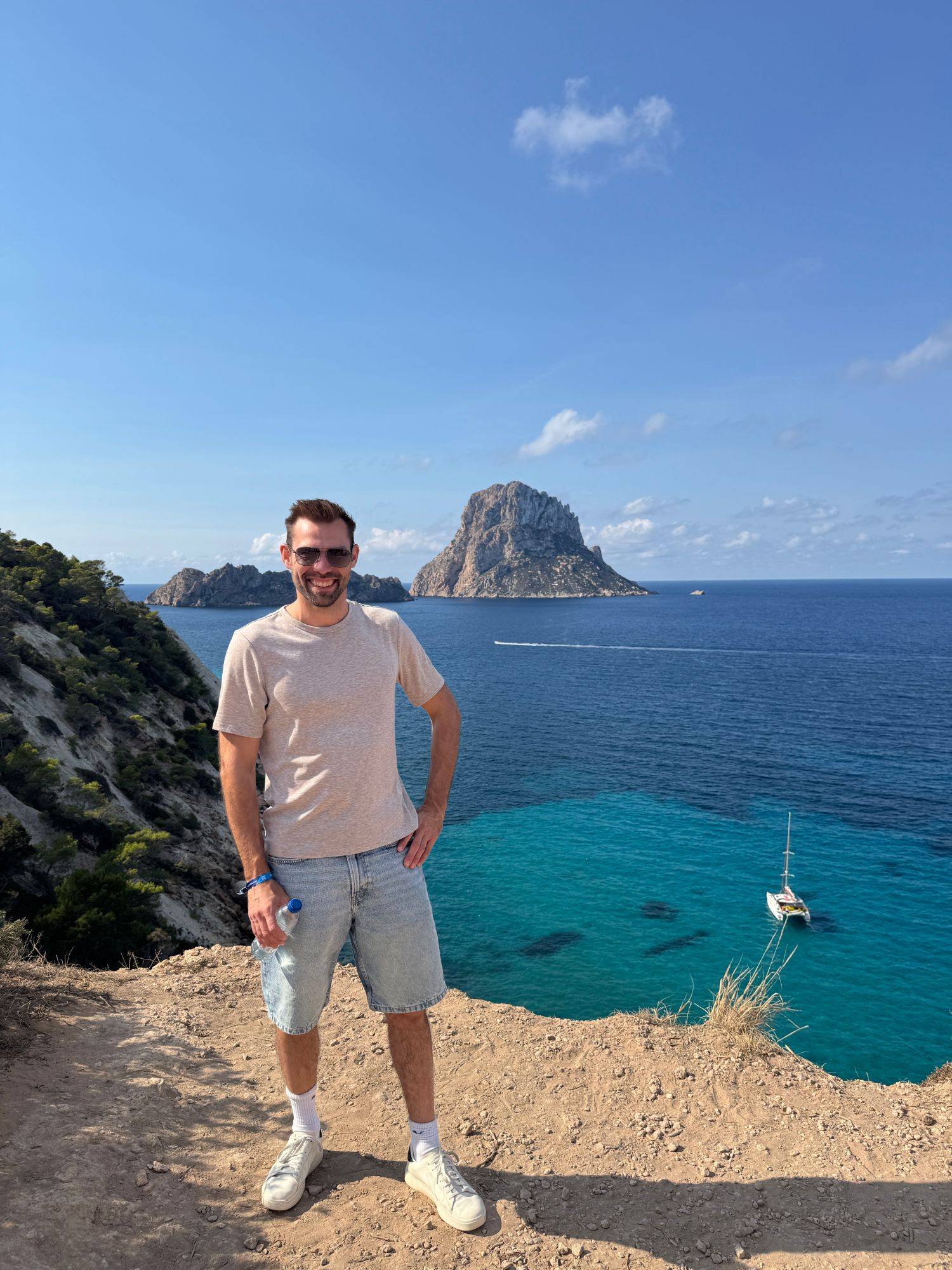 Edwin smiling on a cliff top above turquoise water, with the rock island Es Vedrà behind him.